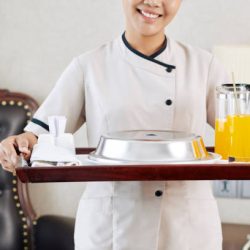 Close-up of young waitress in uniform holding tray with breakfast and juice and smiling while standing in the hotel room